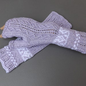 May include: A pair of lavender fingerless gloves with intricate knitted patterns. The gloves feature openwork designs and white snowflake-like accents on the cuffs and upper hand. The gloves are made of a soft, textured yarn.