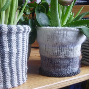 以下が含まれることがあります： 異なるパターンと色のニット製の植木鉢が3つあります。鉢には、緑色の多肉植物やアロエベラなど、さまざまな植物が植えられています。