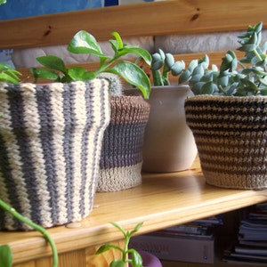 以下が含まれることがあります： 木製の棚に置かれた鉢植えの植物のコレクション。鉢は、ニットやセラミックのデザインなど、さまざまなスタイルです。ニットの鉢は、ニュートラルカラーのストライプ模様が特徴です。さまざまな葉の形をした緑の植物が鉢を満たし、自然な美しさを生み出しています。