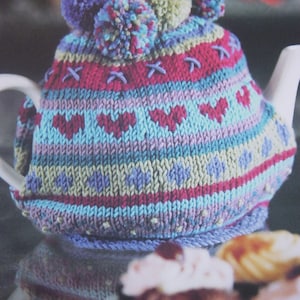 Könnte beinhalten: Ein farbenfroher, gestrickter Teewärmer mit Bommeln oben. Der Wärmer hat ein Muster aus Herzen, Kreuzen und geometrischen Formen in Blau-, Rosa-, Lila- und Grüntönen. Der Wärmer ist für eine weiße Teekanne konzipiert.