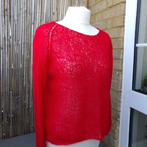 May include: A red knitted jumper with a loose fit and a round neckline. The jumper is made of a lightweight, airy fabric.