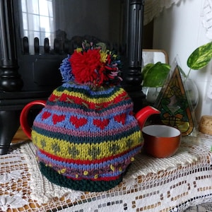 May include: A red teapot with a colorful, knitted tea cozy featuring red hearts and a large pom-pom. A small orange teacup sits nearby on a lace doily. The cozy has blue, yellow, and green stripes.