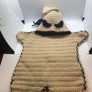 May include: A tan crochet baby costume resembling a character with a pointed hood, black eyes, and a black stitched outline. The costume has a star-like shape with a scalloped edge and a black mustache detail.