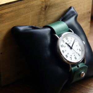May include: A silver wristwatch with a white face and black hands. The watch has a green leather band and is displayed on a black leather watch pillow.
