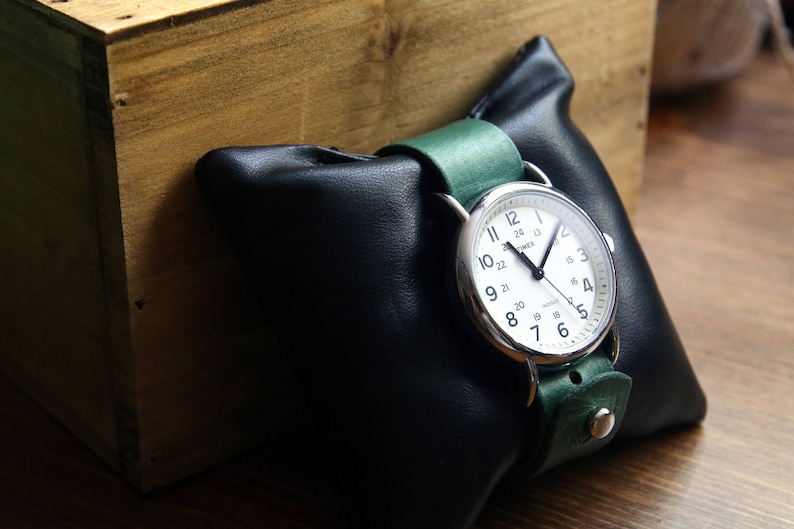 May include: A silver wristwatch with a white face and black hands. The watch has a green leather band and is displayed on a black leather watch pillow.