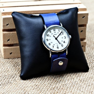 May include: A silver Timex watch with a white face and black hands. The watch has a blue leather band and is displayed on a black pillow.