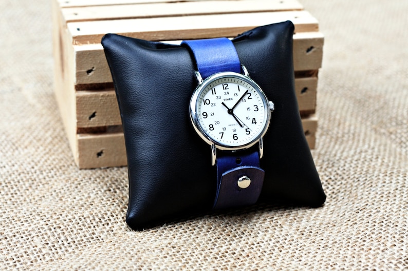 May include: A silver Timex watch with a white face and black hands. The watch has a blue leather band and is displayed on a black pillow.