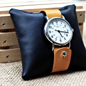 May include: A silver watch with a white face and black hands on a tan leather band. The watch is sitting on a black leather watch pillow.