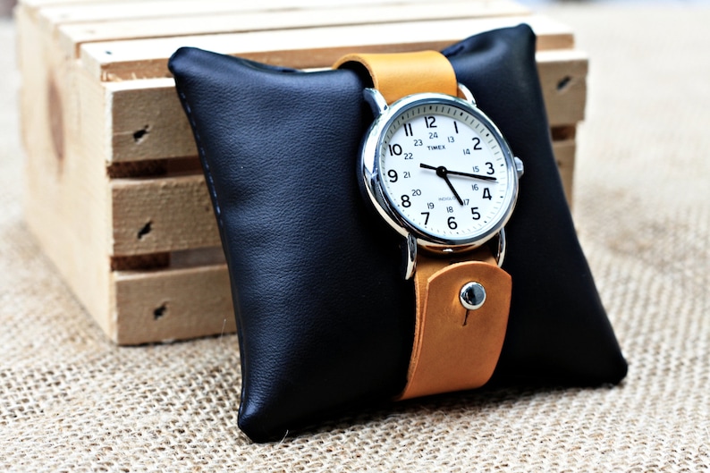 May include: A silver watch with a white face and black hands on a tan leather band. The watch is sitting on a black leather watch pillow.