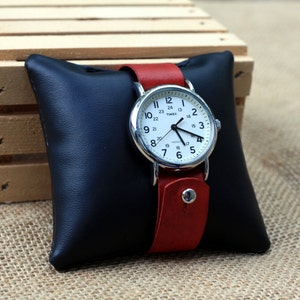 May include: A silver wristwatch with a white face and black numbers. The watch has a red leather band and is displayed on a black pillow.