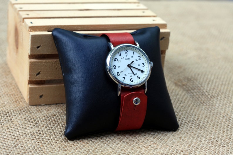 May include: A silver wristwatch with a white face and black numbers. The watch has a red leather band and is displayed on a black pillow.