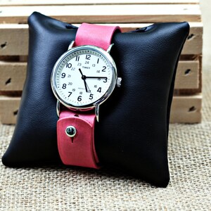 May include: A silver wristwatch with a white face and black hands. The watch has a pink leather band and is displayed on a black leather cushion.