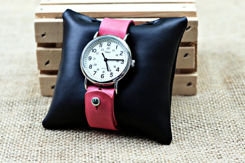 May include: A silver wristwatch with a white face and black hands. The watch has a pink leather band and is displayed on a black leather cushion.