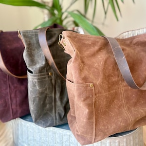 May include: Three waxed canvas tote bags in shades of burgundy, olive green, and brown. Each bag has a leather strap and an exterior pocket. The bags are sitting on a metal surface, with a green plant in the background.
