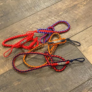 May include: Three braided paracord lanyards featuring metal identification tags. One lanyard is solid red, one is a combination of blue and red, and one is a combination of orange and dark blue. Each cord includes a loop end and a metal plate.