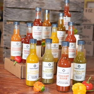 May include: A collection of 12 small glass bottles of hot sauce with various flavors, including Jalapeno Lime, Cherry Bomb, Fresh Tomatillo, and Fatali Attraction. The bottles are arranged in two rows on a wooden surface with a brown box in front of them.  The bottles have black caps and white labels with the brand name "Bushel & Peck's" printed on them.