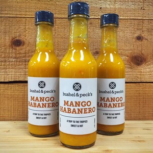 May include: Three glass bottles of mango habanero hot sauce with black caps. The labels are white with black text that reads "bushel & peck's Mango Habanero A Trip to the Tropics Sweet & Hot".