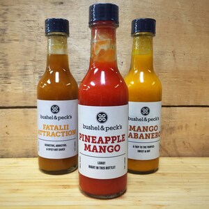 May include: Three glass bottles of hot sauce with black caps and white labels. The labels read "Fatalii Attraction", "Pineapple Mango", and "Mango Habanero". The sauces are various shades of orange and red. The background is a wooden surface.
