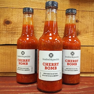 May include: Three glass bottles of cherry bomb hot sauce with a red and white label. The label reads "bushel & peck's Cherry Bomb Highly Addictive Hot Sauce with a Pop of Heat & Smokiness".