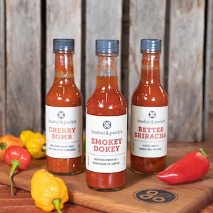 May include: Three glass bottles of hot sauce with black caps and labels. The labels are white with black text and a logo. The bottles are arranged in a row on a wooden surface. The hot sauces are labeled "Cherry Bomb", "Smokey Dokey", and "Better Sriracha".