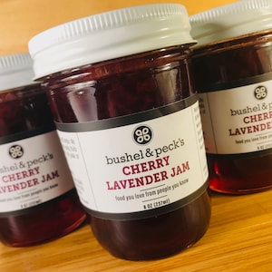 May include: Three glass jars of cherry lavender jam with white lids. The labels are black with white text that reads "bushel & peck's CHERRY LAVENDER JAM food you love from people you know 8 oz (237ml)"