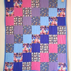 May include: A colourful patchwork quilt with a blue border, featuring a grid of squares in pink, purple, and blue. Some squares have cartoon and space-themed patterns. The quilt is hung with wooden pegs. The quilt measures approximately 100 cm x 100 cm.