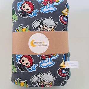 May include: A folded grey fabric blanket featuring a repeating cartoon pattern of superheroes including Thor, Iron Man, and Black Widow. A brown paper band wraps around the centre with a circular sticker reading moon & mallow, plus a small white tag.