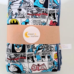 May include: A folded fabric blanket featuring a vintage Batman comic book print with black, white, blue, and red panels. A tan paper band wraps around the centre, displaying a circular white sticker with the text moon & mallow in black and yellow.