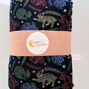 May include: A black fabric item featuring a repeating pattern of colourful magical house crests, including a green snake, red lion, blue eagle, and yellow badger, with white stars. A tan paper band with a moon and mallow logo wraps around the centre.