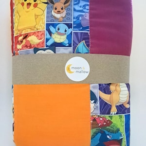 May include: A folded blanket with panels displaying Pokémon characters like Pikachu and Eevee. The blanket is wrapped with a brown band and a sticker with the text "moon & mallow". The blanket has orange and purple sections.