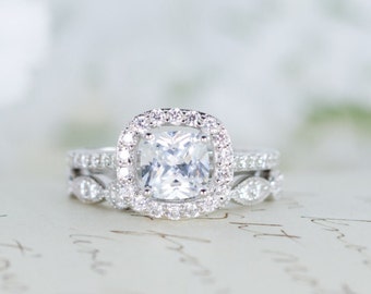 Op de afbeelding: Een sprankelende verlovingsring met een grote, vierkant geslepen middensteen omringd door een halo van kleinere diamanten. De band is ook versierd met diamanten, wat een schitterende weergave creëert. De ring ligt op een oppervlak met handschrift.