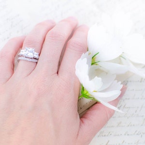 May include: A close-up of a hand wearing a silver ring with a large, clear, square-shaped diamond and two smaller diamonds on either side. The hand is resting on a white background with white flowers.