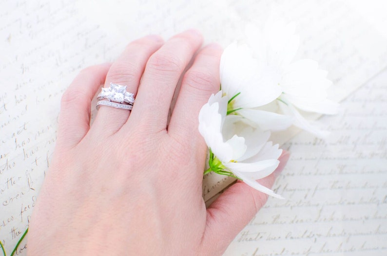 May include: A close-up of a hand wearing a silver ring with a large, clear, square-shaped diamond and two smaller diamonds on either side. The hand is resting on a white background with white flowers.