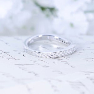 May include: A silver wedding band with a delicate, leaf-shaped design. The band is set with small, clear stones.