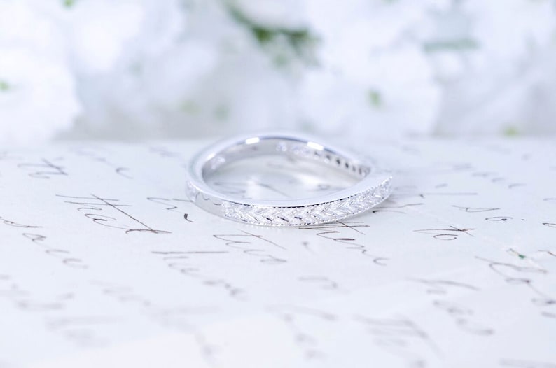 May include: A silver wedding band with a delicate, leaf-shaped design. The band is set with small, clear stones.