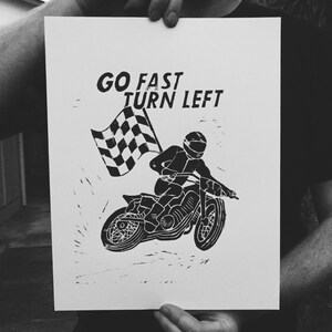 May include: Black and white print featuring a motorcycle racer holding a checkered flag. The text "GO FAST TURN LEFT" is above the racer. The image is a linocut style illustration.