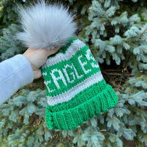 May include: A green and white knit beanie with the word "EAGLES" in white letters. The hat has a gray faux fur pom-pom on top. The hat is held up in front of a background of green pine needles.