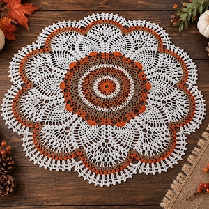 May include: A round, intricate crochet doily featuring a central pattern in shades of burnt orange and brown, transitioning into a wide, scalloped white lace border with delicate openwork stitching and small orange and brown accents throughout.