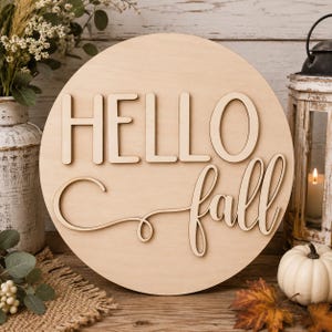May include: A circular light-colored wooden sign featuring the word HELLO in a bold, sans-serif font and the word fall in a cursive script. The text is raised and mounted on a flat, round wooden board with a smooth, natural finish.