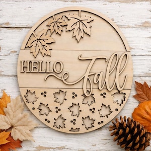 May include: A circular light-colored wooden sign featuring the laser-cut text "HELLO Fall" in a mix of block and script lettering. The design includes cutout maple leaf shapes at the top and a pattern of small leaves and berries at the bottom.