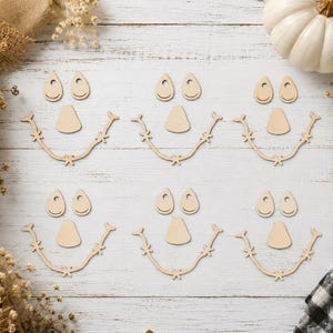 May include: Six sets of unfinished wooden craft pieces, each containing two teardrop-shaped eyes, one triangular nose, and one curved mouth with stitched-style detailing. These light-colored wooden cutouts are arranged in two rows of three.