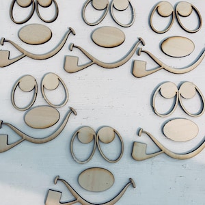 May include: Wooden cutouts of cartoon faces, including eyes, noses, and smiles. The pieces are unfinished, with a light brown color, and are arranged on a white surface. The shapes are designed for crafting or DIY projects.