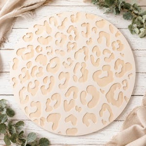 May include: A circular light-colored wooden sign featuring a laser-cut leopard print pattern. The design consists of various irregular, organic shapes cut out from the flat, round wooden surface, creating a repeating animal-inspired motif.