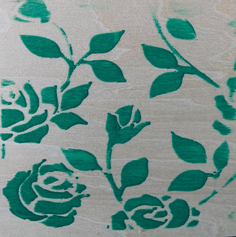 May include: A light-colored wooden surface featuring a painted pattern of vibrant green roses and leaves. The design consists of multiple rose blossoms in various stages of bloom, connected by stems and foliage, applied directly onto the wood grain.