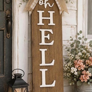 May include: A vertical wooden sign featuring the white cutout text "oh HELLO" arranged vertically. The sign is adorned at the top with a large beige and white checkered burlap bow and artificial greenery. The text "OH HELLO CUTOUTS ONLY BOARD NOT INCLUDED" is printed at the bottom.