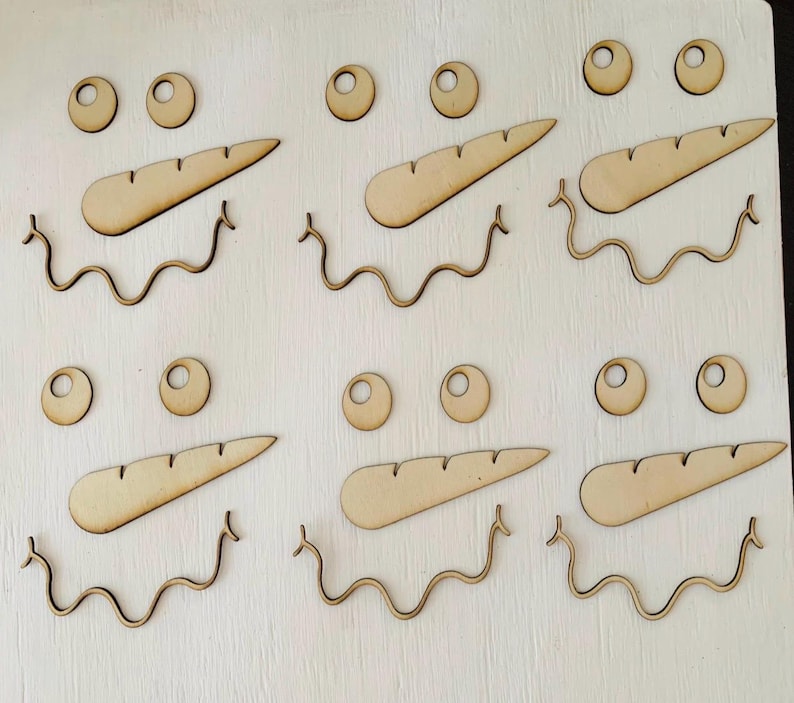 May include: Six sets of laser-cut wooden snowman face components arranged in two rows. Each set includes two circular eyes with centre holes, one elongated carrot-shaped nose with etched lines, and one wavy line representing a smiling mouth.