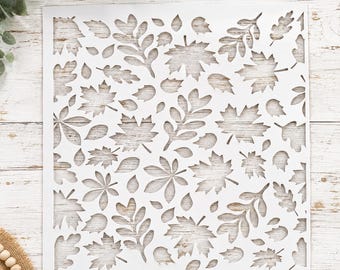 May include: A square white stencil featuring a repeating cutout pattern of various autumn leaves, including maple, oak, and fern shapes. The design covers the entire surface of the stencil, creating a dense arrangement of foliage silhouettes.