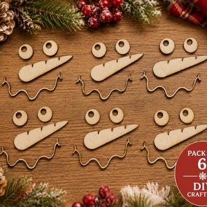 May include: A set of six wooden snowman face craft kits, each containing two circular eyes, one carrot-shaped nose, and one wavy mouth piece. A red circular badge in the corner displays the text PACK OF 6 DIY CRAFT KIT.
