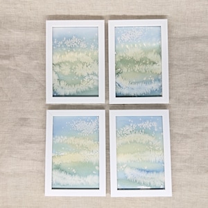May include: Four framed watercolour paintings in white frames. Each painting depicts a landscape with blue, green, and yellow hues, and white textured details. The artwork is displayed on a neutral-coloured surface.