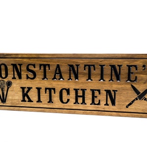 以下が含まれることがあります： 黒い文字で「CONSTANTINE'S KITCHEN」と書かれた木製の看板。看板は濃い茶色に着色され、装飾的な縁取りが施されています。木にはキッチン用品と交差したナイフが彫刻されています。